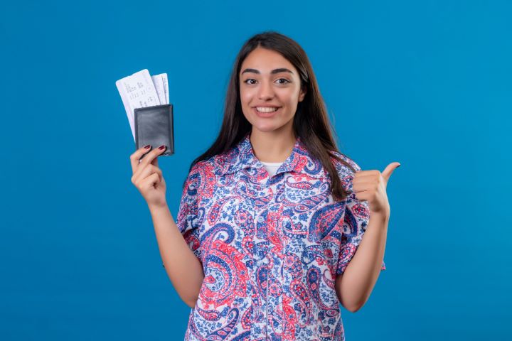 young-beautiful-woman-tourist-holding-passport-with-tickets-smiling-cheerfully-showing-thumbs-up-ready-holiday-isolated-blue-wall (Small)